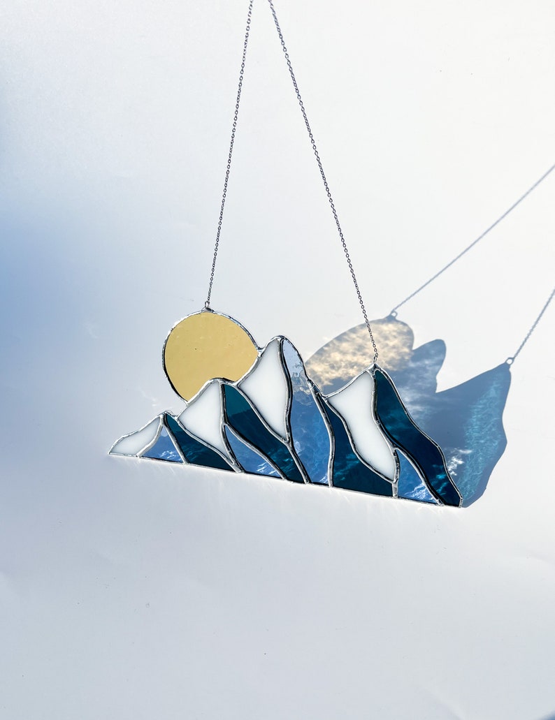 May include: A stained glass sun and mountain scene hanging from a silver chain. The sun is yellow, the mountains are white and blue, and the background is a light blue.