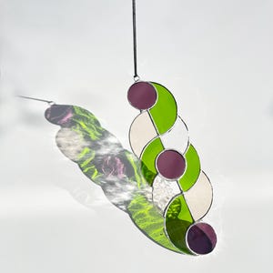 Puede incluir: Un atrapasoles de vidriera con un dise&ntilde;o geom&eacute;trico. La pieza presenta c&iacute;rculos entrelazados en tonos de verde, blanco y morado, con un cord&oacute;n negro para colgar. El atrapasoles proyecta una sombra colorida sobre el fondo blanco.