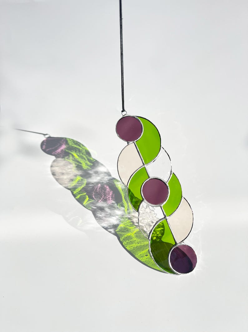 Puede incluir: Un atrapasoles de vidriera con un dise&ntilde;o geom&eacute;trico. La pieza presenta c&iacute;rculos entrelazados en tonos de verde, blanco y morado, con un cord&oacute;n negro para colgar. El atrapasoles proyecta una sombra colorida sobre el fondo blanco.