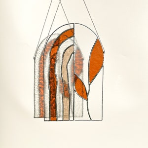 May include: Stained glass window hanging with an arched top. The design features orange, tan, and clear glass panels, with a leaf-like shape on the right side. The piece is suspended by a silver chain.