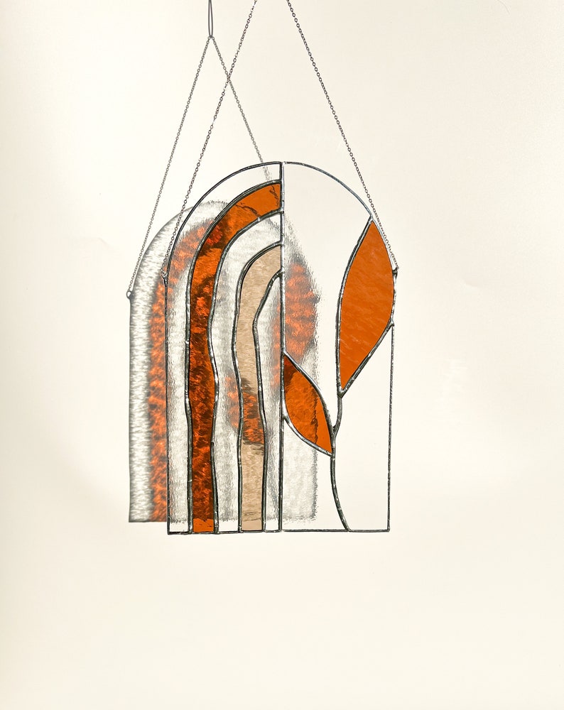 May include: Stained glass window hanging with an arched top. The design features orange, tan, and clear glass panels, with a leaf-like shape on the right side. The piece is suspended by a silver chain.