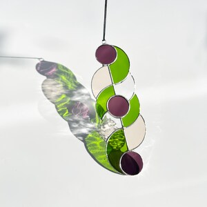 Puede incluir: Atrapasoles de vidriera con un dise&ntilde;o &uacute;nico. La pieza presenta una serie de formas conectadas en verde, blanco y morado. Est&aacute; suspendido por un cord&oacute;n negro, proyectando una sombra colorida sobre un fondo blanco.