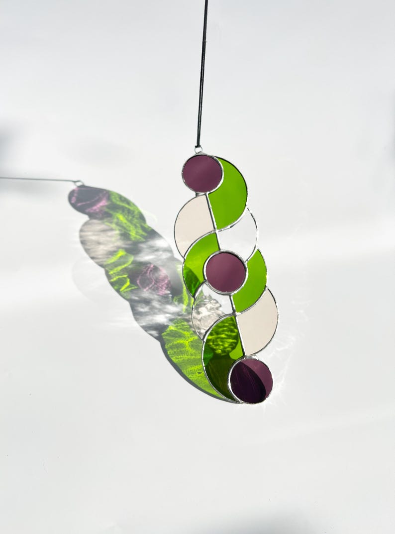 Puede incluir: Atrapasoles de vidriera con un dise&ntilde;o &uacute;nico. La pieza presenta una serie de formas conectadas en verde, blanco y morado. Est&aacute; suspendido por un cord&oacute;n negro, proyectando una sombra colorida sobre un fondo blanco.