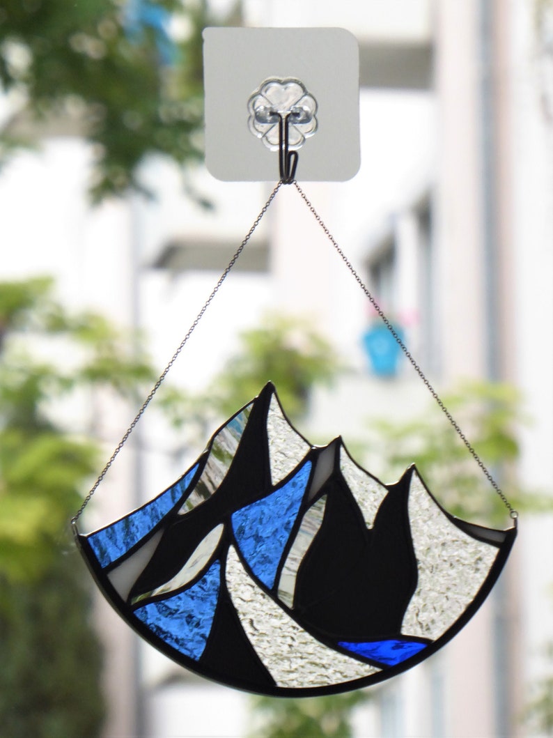 Pu&ograve; includere: Un acchiappasole in vetro colorato con un design a catena montuosa, composto da segmenti di vetro blu, bianco e nero testurizzati uniti da linee di saldatura in metallo scuro, appeso a una sottile catena metallica fissata a un gancio adesivo.