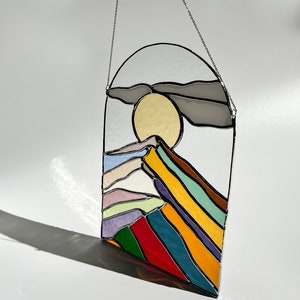 May include: A stained glass window hanging from a silver chain. The window depicts a stylized mountain range with a yellow sun and a gray cloud. The mountain range is made up of colorful stripes in shades of purple, blue, green, yellow, and red.