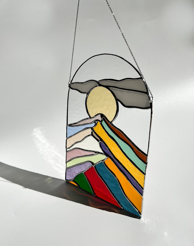 May include: A stained glass window hanging from a silver chain. The window depicts a stylized mountain range with a yellow sun and a gray cloud. The mountain range is made up of colorful stripes in shades of purple, blue, green, yellow, and red.