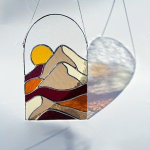 K&ouml;nnte beinhalten: Ein Buntglasfenster, das an einer Kette h&auml;ngt. Das Fenster zeigt einen Sonnenuntergang &uuml;ber einem Gebirge mit einer gelben Sonne, braunen Bergen und roten, orangen und cremefarbenen Schichten.