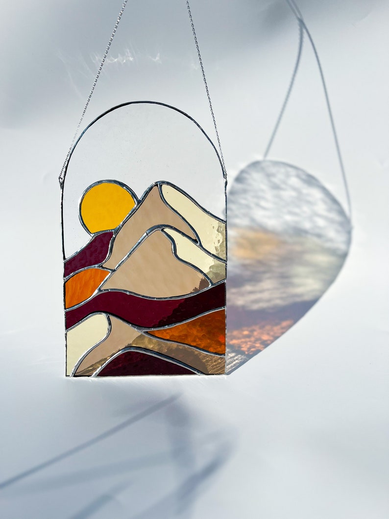 K&ouml;nnte beinhalten: Ein Buntglasfenster, das an einer Kette h&auml;ngt. Das Fenster zeigt einen Sonnenuntergang &uuml;ber einem Gebirge mit einer gelben Sonne, braunen Bergen und roten, orangen und cremefarbenen Schichten.