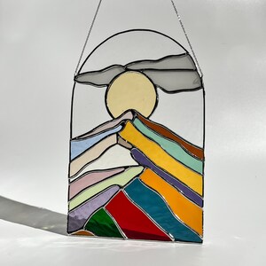 May include: A stained glass window hanging with a chain. The window depicts a stylized mountain range with a yellow sun and a gray cloud above. The mountains are made of different colored glass, including red, green, blue, yellow, and purple.