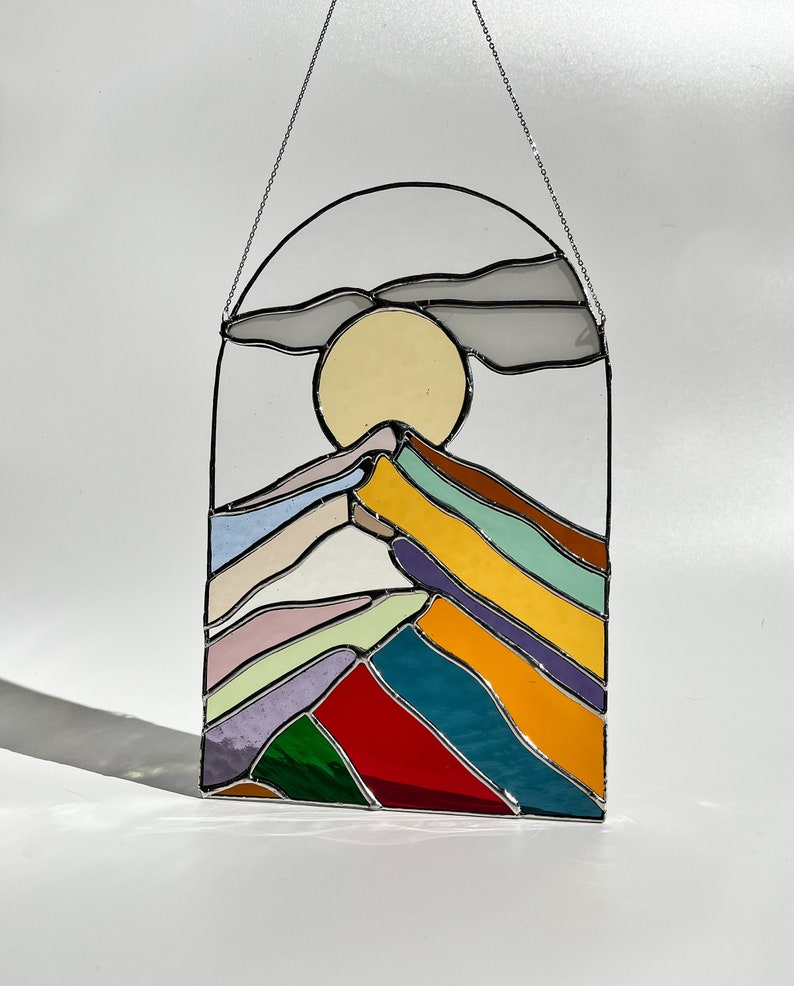 May include: A stained glass window hanging with a chain. The window depicts a stylized mountain range with a yellow sun and a gray cloud above. The mountains are made of different colored glass, including red, green, blue, yellow, and purple.