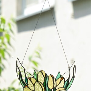 May include: A stained glass tulip design, hanging from a chain. The piece features yellow and green glass, with a crescent shape. The tulips are clustered together, with green leaves. The piece is suspended from a clear adhesive hook.