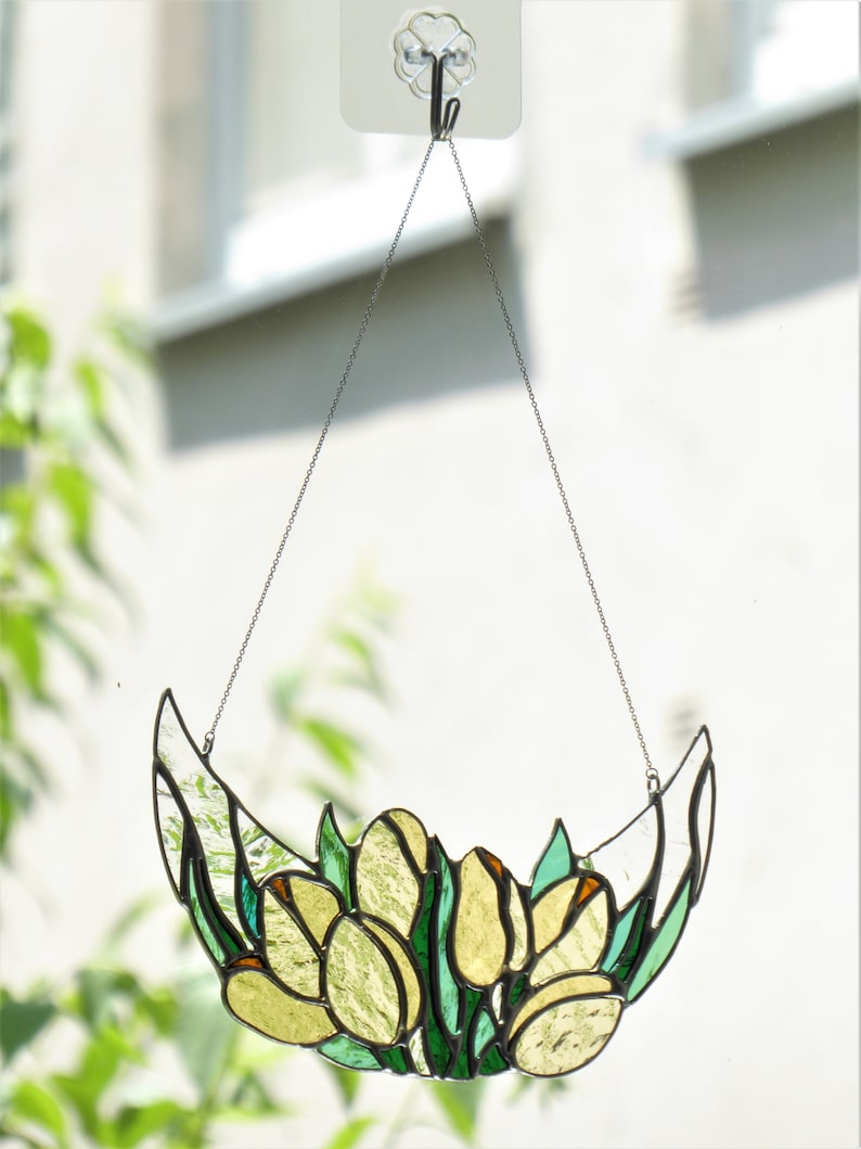 May include: A stained glass tulip design, hanging from a chain. The piece features yellow and green glass, with a crescent shape. The tulips are clustered together, with green leaves. The piece is suspended from a clear adhesive hook.