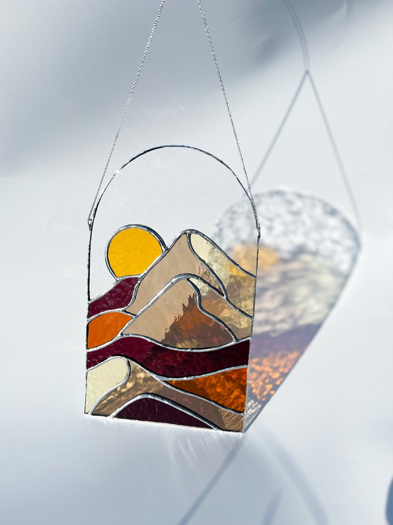 K&ouml;nnte beinhalten: Eine Sonne und eine Berglandschaft aus Buntglas, die an einer silbernen Kette h&auml;ngt. Die Sonne ist gelb, die Berge sind braun und der Hintergrund ist eine Mischung aus Orange, Rot und Creme.