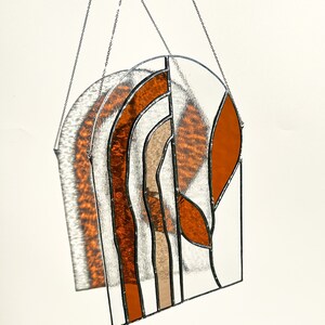 May include: Stained glass suncatcher with a geometric design. The piece features orange, tan, and clear glass sections, framed with black lines and suspended by a silver chain. The suncatcher is arched at the top and casts a shadow.