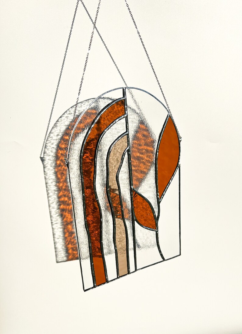 May include: Stained glass suncatcher with a geometric design. The piece features orange, tan, and clear glass sections, framed with black lines and suspended by a silver chain. The suncatcher is arched at the top and casts a shadow.