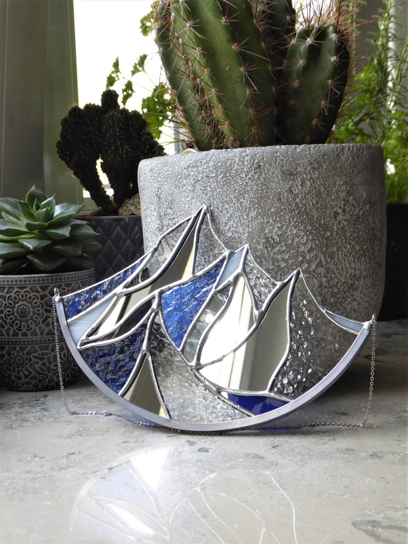 Pu&ograve; includere: Un acchiappasole in vetro colorato a forma di semicerchio con un design a catena montuosa. L'oggetto &egrave; composto da pannelli di vetro blu testurizzato, vetro a specchio trasparente e vetro iridescente, uniti da linee di saldatura argentate.