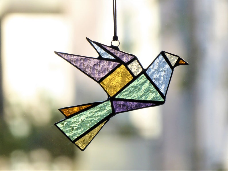 Pode incluir: Um apanhador de sol de p&aacute;ssaro origami em vitral em tons de roxo, amarelo, verde e azul. O design geom&eacute;trico apresenta um contorno preto e &eacute; suspenso por um cord&atilde;o preto. O p&aacute;ssaro &eacute; um item decorativo.