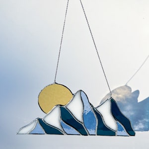 May include: A stained glass sun and mountain scene hanging from a silver chain. The sun is yellow, the mountains are blue and white, and the background is a light blue.