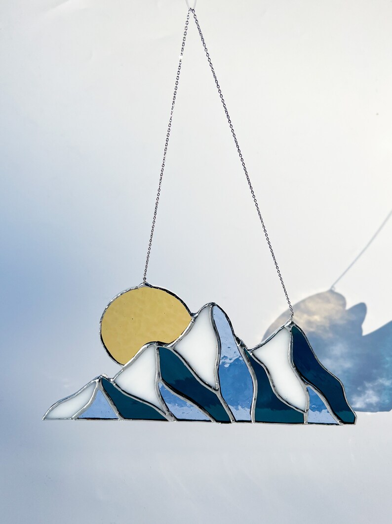 May include: A stained glass sun and mountain scene hanging from a silver chain. The sun is yellow, the mountains are blue and white, and the background is a light blue.