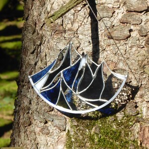 Pu&ograve; includere: Un acchiappasole in vetro colorato a forma di semicerchio con un design a catena montuosa. L'oggetto &egrave; composto da singoli pannelli di vetro blu scuro, nero e trasparente tenuti insieme da linee di saldatura in metallo argentato e una catena.