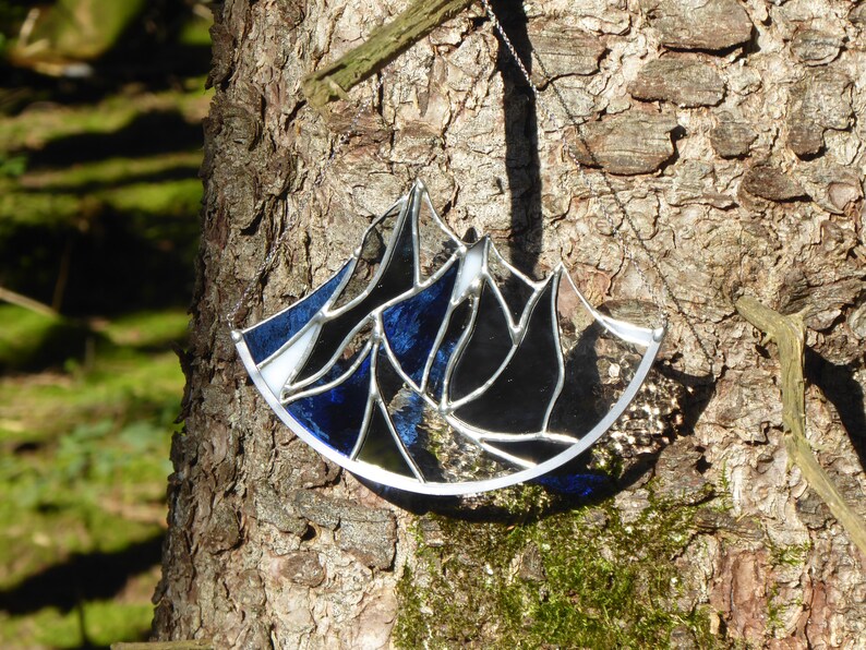 Pu&ograve; includere: Un acchiappasole in vetro colorato a forma di semicerchio con un design a catena montuosa. L'oggetto &egrave; composto da singoli pannelli di vetro blu scuro, nero e trasparente tenuti insieme da linee di saldatura in metallo argentato e una catena.