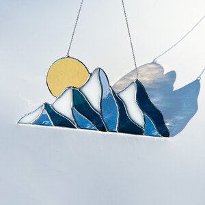 May include: A stained glass sun and mountain scene hanging from a silver chain. The sun is yellow and the mountains are blue and white.
