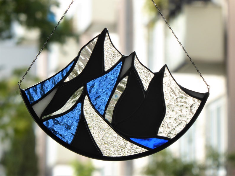 Pu&ograve; includere: Un acchiappasole in vetro colorato a forma di semicerchio con un design a catena montuosa. L'oggetto &egrave; composto da segmenti di vetro trasparente testurizzato, vetro nero solido e vetro blu vibrante uniti da linee di saldatura in metallo scuro.