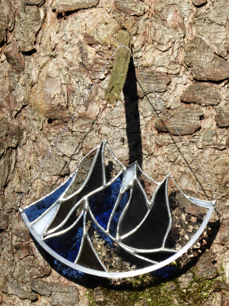 Pu&ograve; includere: Un acchiappasole in vetro colorato a forma di semicerchio con un design a catena montuosa. L'oggetto &egrave; composto da segmenti di vetro nero, blu scuro e bianco uniti da linee di saldatura in metallo argentato e include una sottile catena.