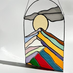 May include: A stained glass window hanging with a colorful mountain scene. The mountain is made of layered glass in shades of purple, green, yellow, red, blue, and white. The sun is a yellow circle behind the mountain. A gray cloud is above the sun.