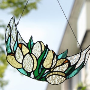 May include: Stained glass crescent moon with floral design. The artwork features white, yellow, green, and teal glass pieces, outlined in black. A chain is attached for hanging. The background is a blurred view of a window and foliage.