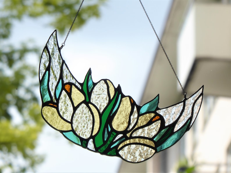 May include: Stained glass crescent moon with floral design. The artwork features white, yellow, green, and teal glass pieces, outlined in black. A chain is attached for hanging. The background is a blurred view of a window and foliage.