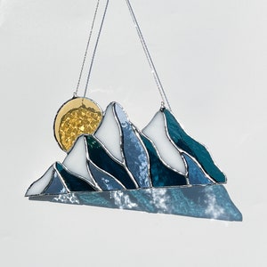 May include: A stained glass sun and mountain scene hanging from a silver chain. The sun is yellow and the mountains are blue and white.