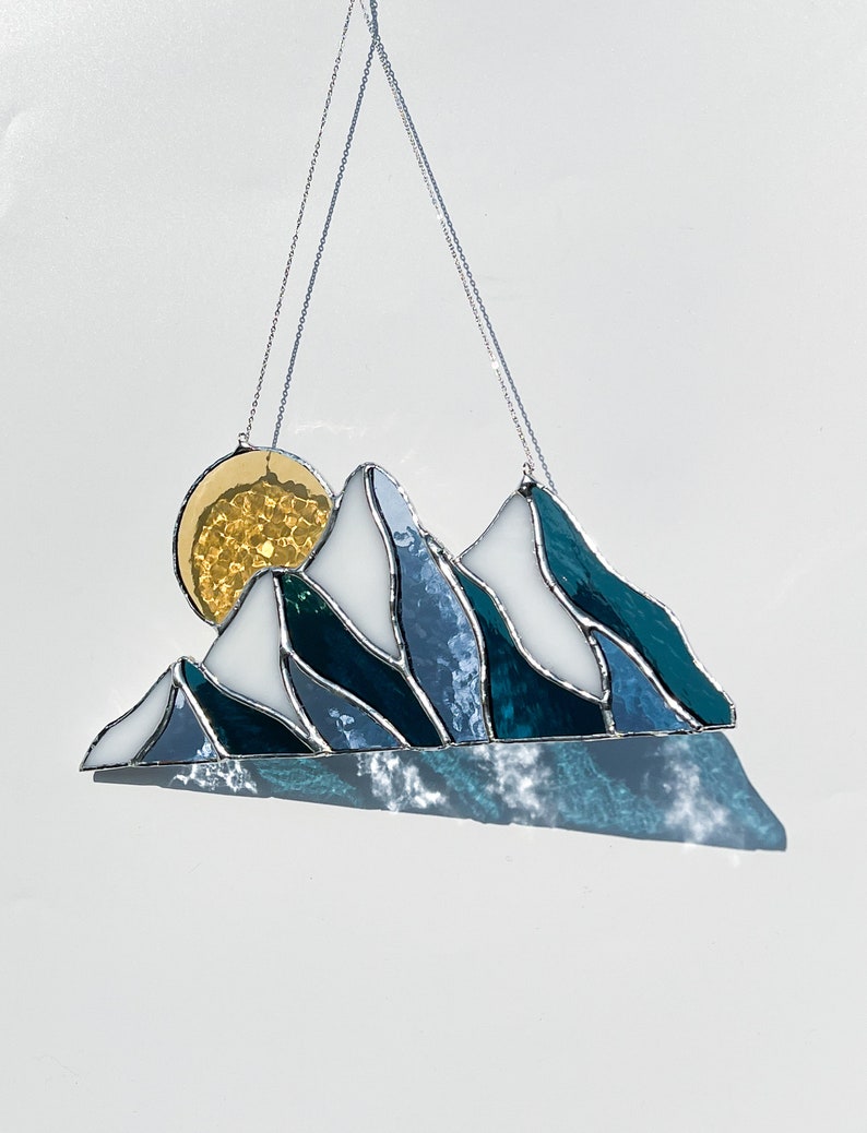 May include: A stained glass sun and mountain scene hanging from a silver chain. The sun is yellow and the mountains are blue and white.
