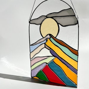 May include: A stained glass window hanging with a colorful mountain scene. The mountain is made of layered glass in shades of purple, green, yellow, red, blue, and white. The sun is a yellow circle behind the mountain. A gray cloud is above the sun.