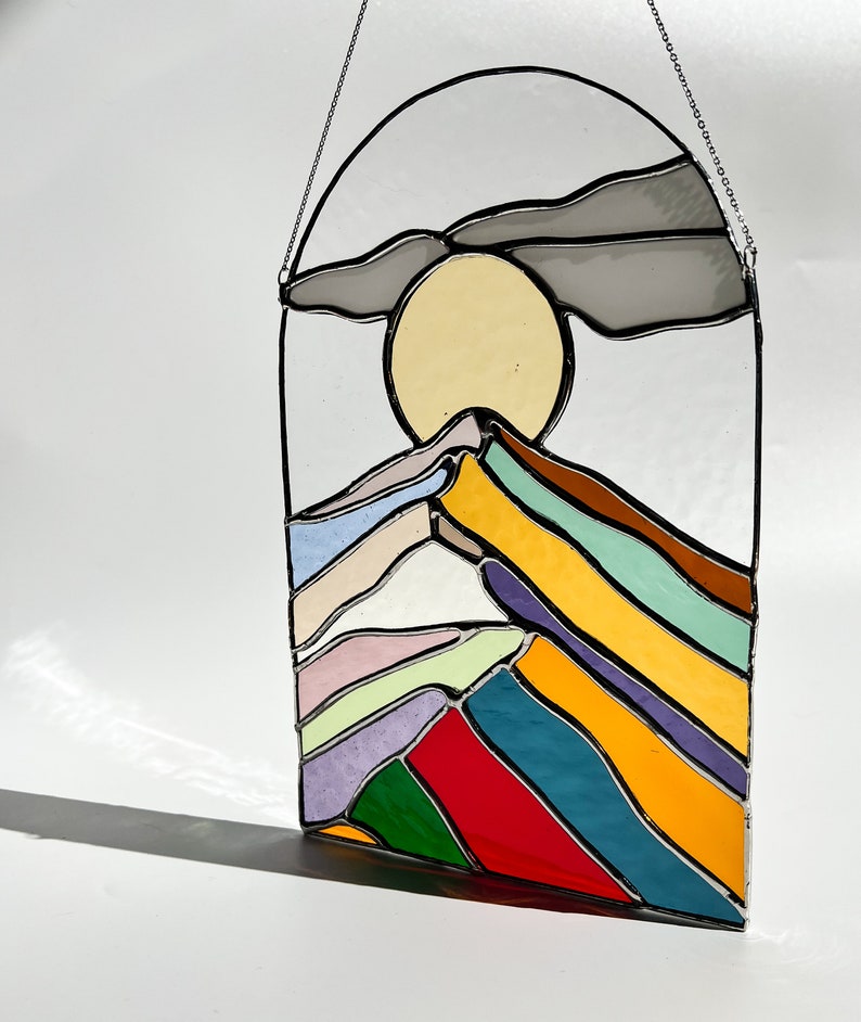 May include: A stained glass window hanging with a colorful mountain scene. The mountain is made of layered glass in shades of purple, green, yellow, red, blue, and white. The sun is a yellow circle behind the mountain. A gray cloud is above the sun.