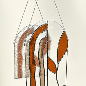 May include: A stained glass window panel with a clear background and orange and tan leaf and arch designs. The panel is suspended by a silver chain. The panel's shadow is visible on the wall behind it.