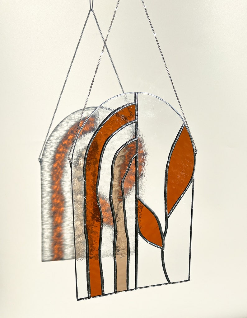 May include: A stained glass window panel with a clear background and orange and tan leaf and arch designs. The panel is suspended by a silver chain. The panel's shadow is visible on the wall behind it.