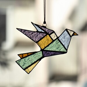 Pode incluir: Um ornamento de p&aacute;ssaro em vitral em estilo origami geom&eacute;trico. O p&aacute;ssaro apresenta uma variedade de cores, incluindo roxo, amarelo, verde e azul, com linhas pretas definindo as formas. Ele pende de um cord&atilde;o preto.