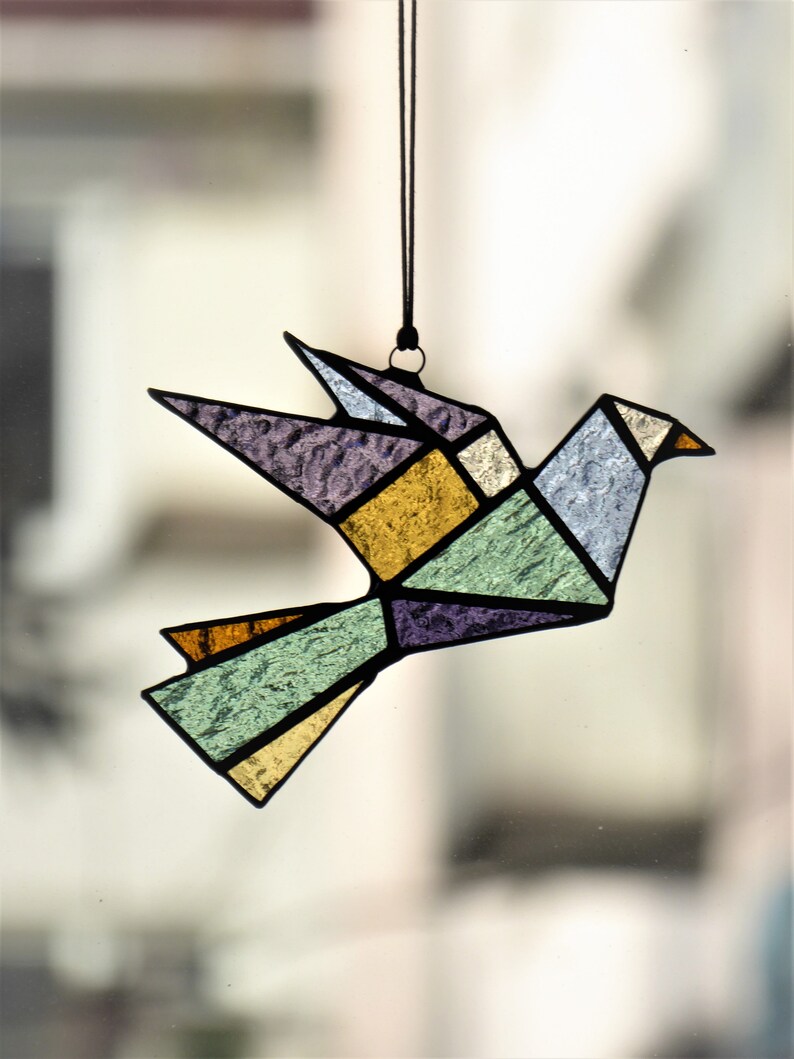 Pode incluir: Um ornamento de p&aacute;ssaro em vitral em estilo origami geom&eacute;trico. O p&aacute;ssaro apresenta uma variedade de cores, incluindo roxo, amarelo, verde e azul, com linhas pretas definindo as formas. Ele pende de um cord&atilde;o preto.
