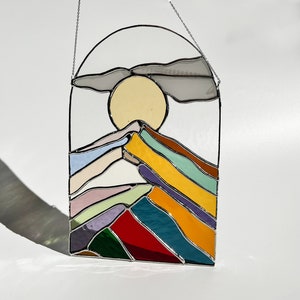 May include: A stained glass window hanging with a chain, depicting a stylized mountain range with a yellow sun and a gray cloud above. The mountains are made of colorful glass in shades of red, orange, yellow, green, blue, purple, and pink.