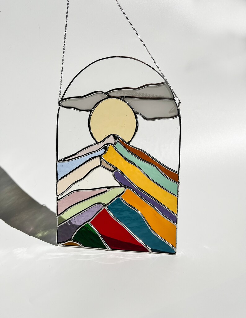 May include: A stained glass window hanging with a chain, depicting a stylized mountain range with a yellow sun and a gray cloud above. The mountains are made of colorful glass in shades of red, orange, yellow, green, blue, purple, and pink.