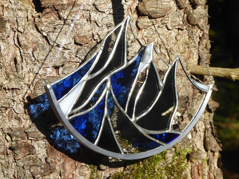 Pu&ograve; includere: Un acchiappasole in vetro colorato con un design a catena montuosa dai picchi frastagliati. L'oggetto &egrave; composto da segmenti di vetro blu traslucido, nero opaco e bianco uniti da linee di saldatura in metallo argentato e include una catenella.