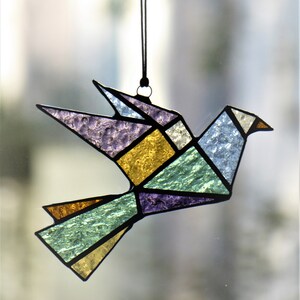 Pode incluir: Um ornamento de p&aacute;ssaro em vitral em estilo origami geom&eacute;trico. O p&aacute;ssaro apresenta v&aacute;rias cores, incluindo roxo, dourado, azul e verde, com contornos pretos. Ele pende de um fio preto, adequado para decora&ccedil;&atilde;o de janelas ou paredes.