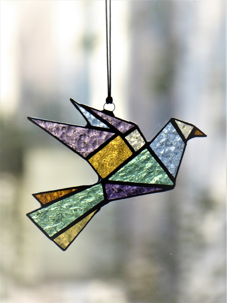 Pode incluir: Um ornamento de p&aacute;ssaro em vitral em estilo origami geom&eacute;trico. O p&aacute;ssaro apresenta v&aacute;rias cores, incluindo roxo, dourado, azul e verde, com contornos pretos. Ele pende de um fio preto, adequado para decora&ccedil;&atilde;o de janelas ou paredes.