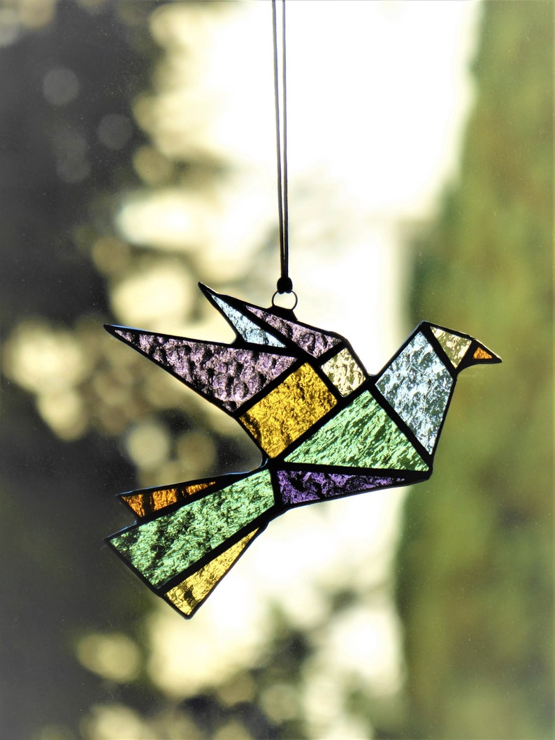 Pode incluir: Apanhador de sol de p&aacute;ssaro em vitral com design geom&eacute;trico de origami. O p&aacute;ssaro apresenta uma variedade de cores, incluindo amarelo, verde, roxo e azul. Ele pendura de um cord&atilde;o preto, perfeito para decora&ccedil;&atilde;o de janelas ou paredes.