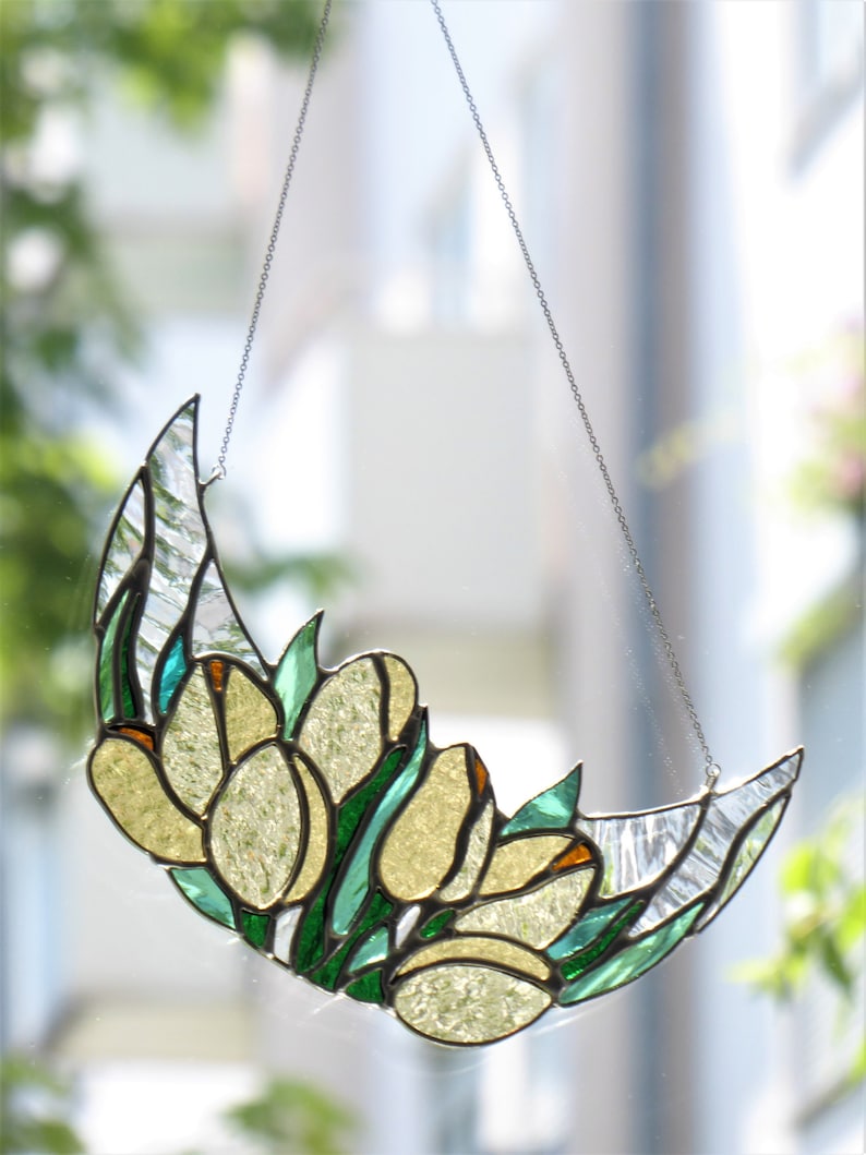 May include: Stained glass crescent moon with yellow tulip design, accented with green leaves and orange tips. The decorative piece hangs from a silver chain. The glass has a textured, translucent appearance, perfect for a window.
