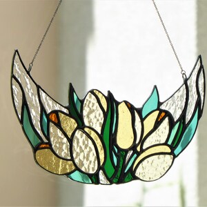 May include: Stained glass floral art piece featuring yellow and white flowers with green leaves, set against a clear background. The piece is crescent-shaped and hangs from a chain. The glass has a textured appearance.