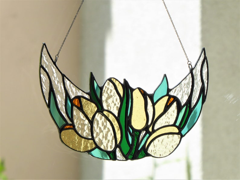 May include: Stained glass floral art piece featuring yellow and white flowers with green leaves, set against a clear background. The piece is crescent-shaped and hangs from a chain. The glass has a textured appearance.
