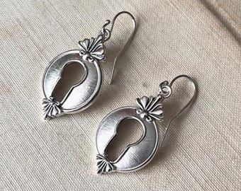 May include: A pair of silver-toned drop earrings featuring a circular frame with a central keyhole cutout. Each earring is suspended from a curved hook wire and displays a small, decorative fan-shaped detail at the top and bottom of the circular frame.