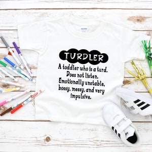 May include: White toddler t-shirt with the word "TURDLER" in a black cloud shape, with a humorous definition below. The image includes colorful markers and white sneakers with black stripes.